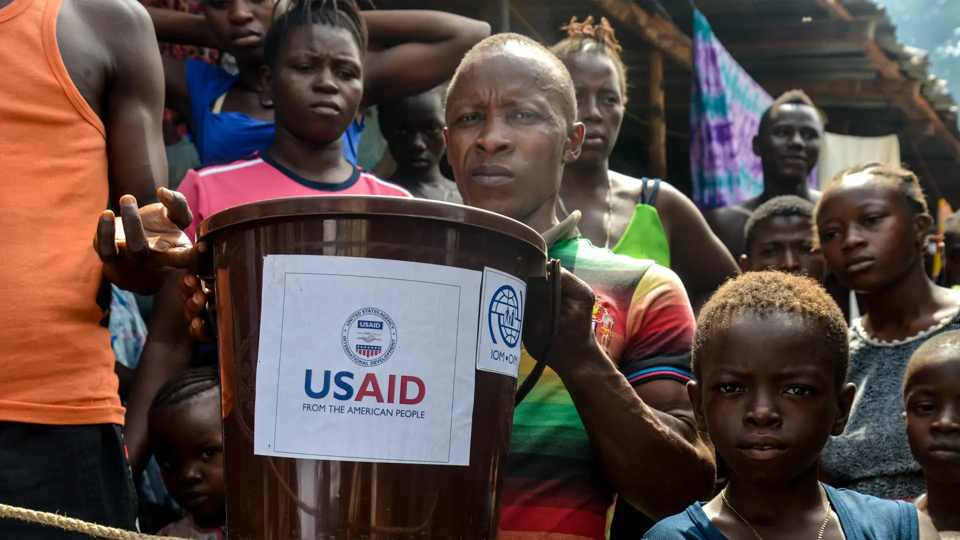 A group of adults and children in Sierra Leone. A man is holding up bucket with US AIid and IOM logos
