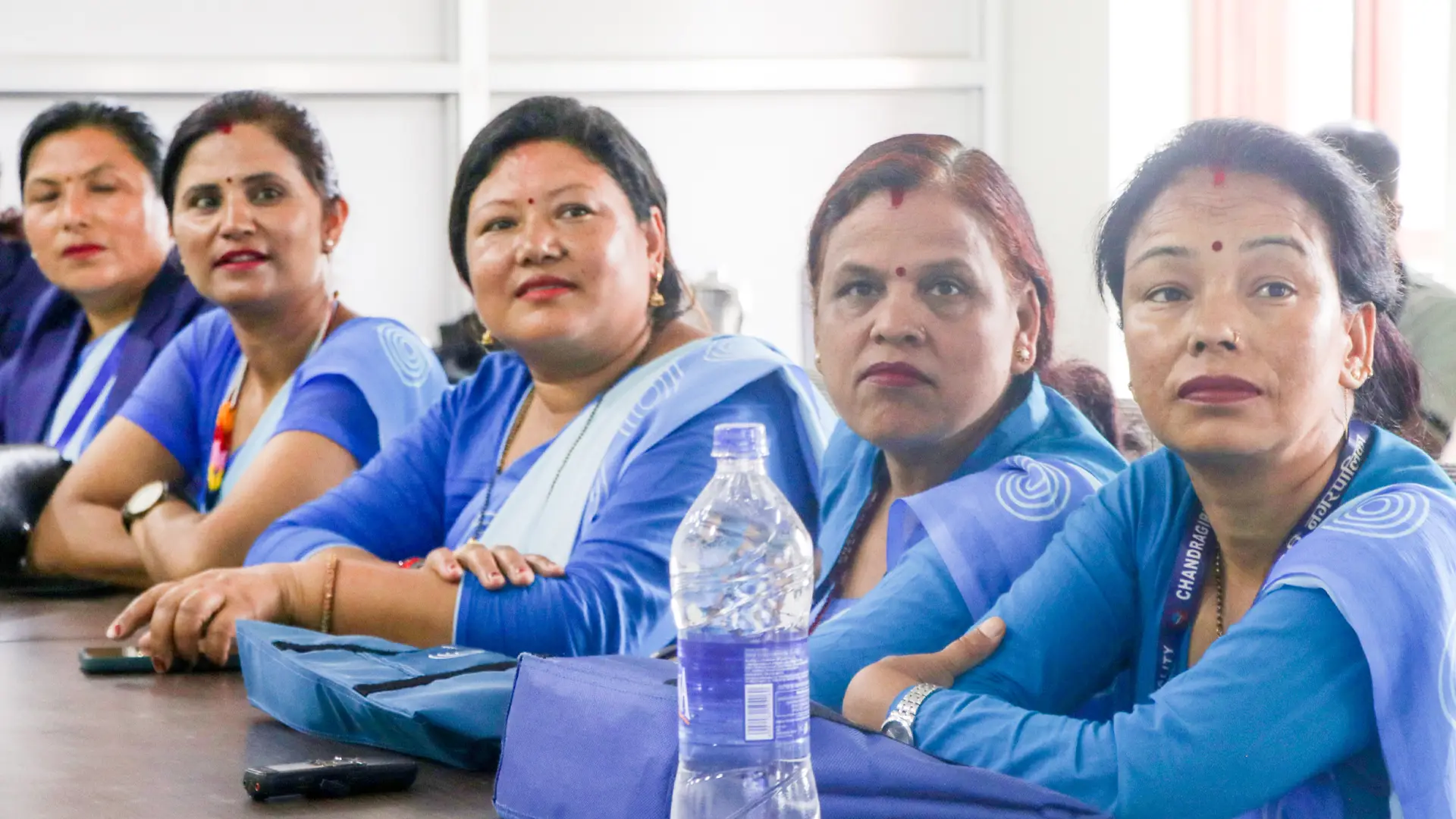 Five seated Nepali women in the same blue sari. They are seated in a row and looking at something behind the viewer