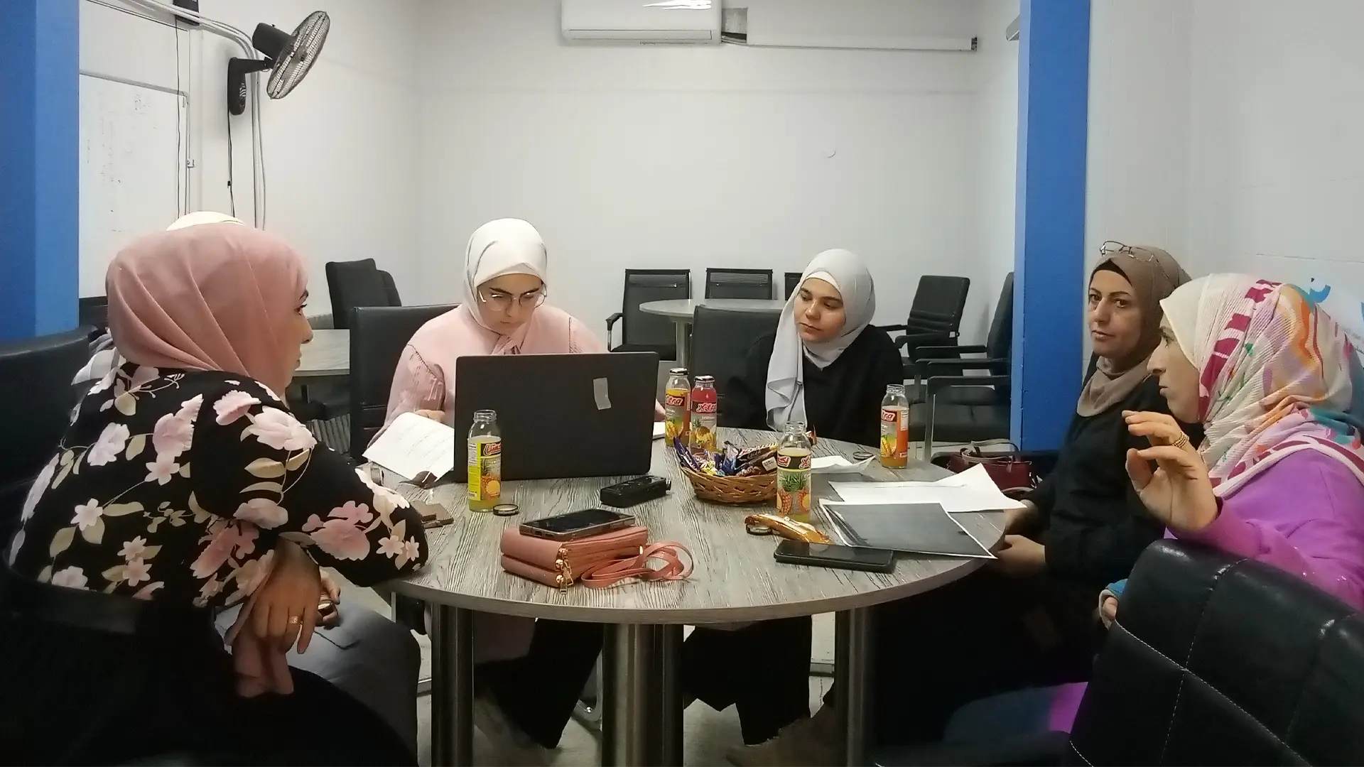 Five Syrian women in headscarves sit around a table talking while one types on a laptop