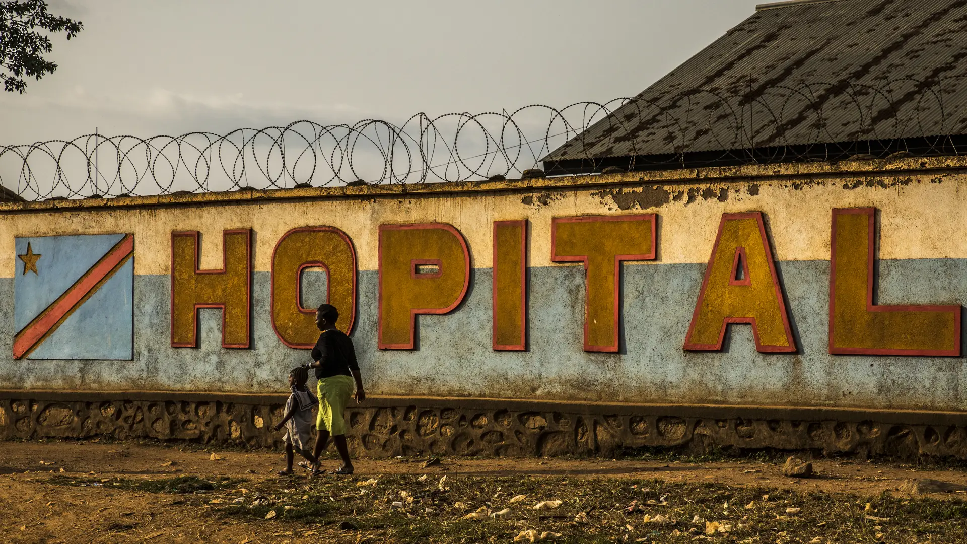 The word 'hopital' on a wall with barbed wire, and an African mother and child walk past