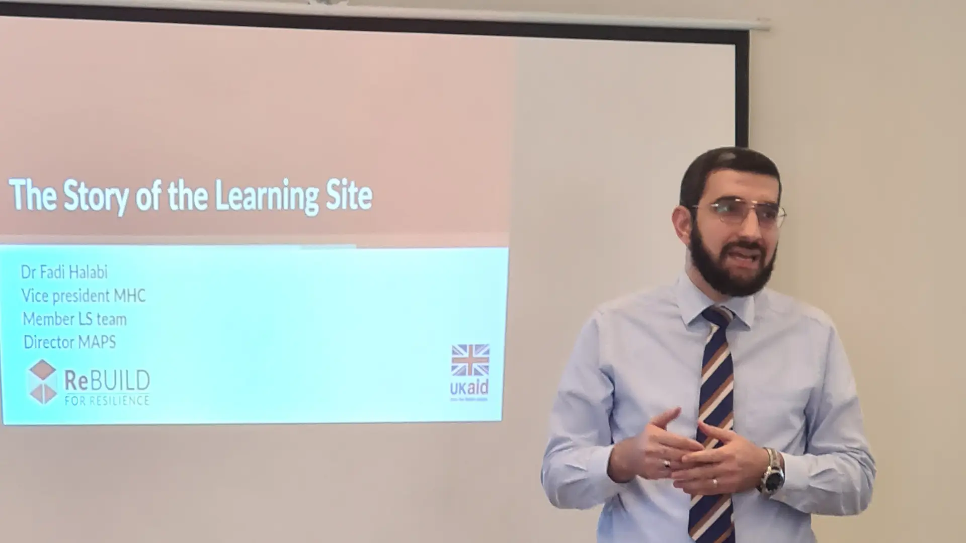 A Syrian man with a beard and glasses stands speaking in front of a screen with the word 'The story of the learning site - Dr Fadi Halabi' and the ReBUILD logo