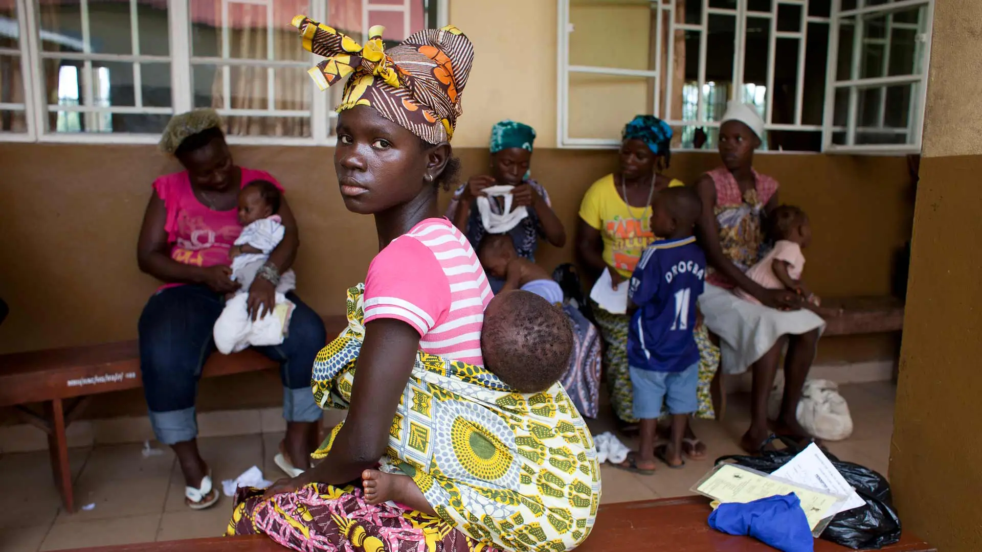 Several Sierra Leonian young women with babies in a waiting room. One woman is turned to look at the viewer