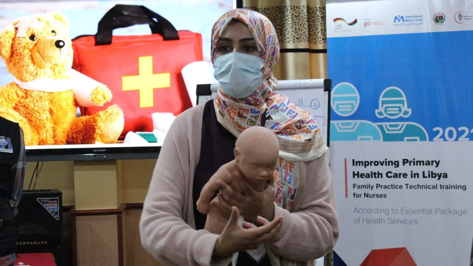 A masked Libyan woman demonstrates how to care for a baby (using a model)