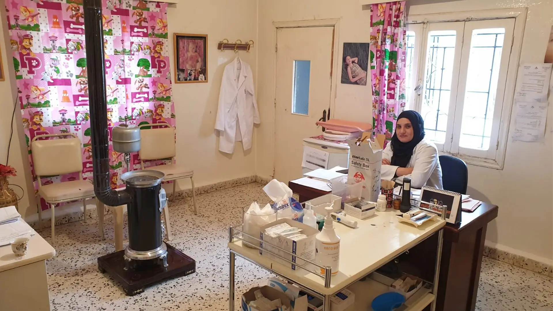 A woman in a black headscarf and white medics coat sits at a desk in a doctor's surgery