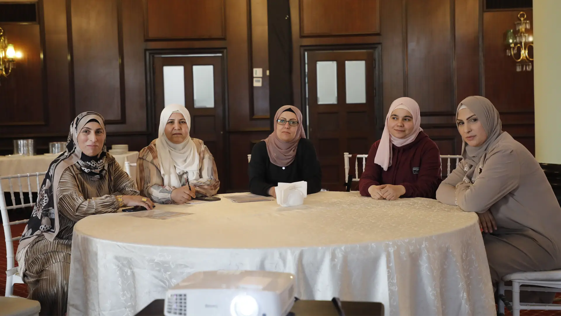 Five Syrian women sit round a table in a wood panelled room, all looking defiantly at the viewer