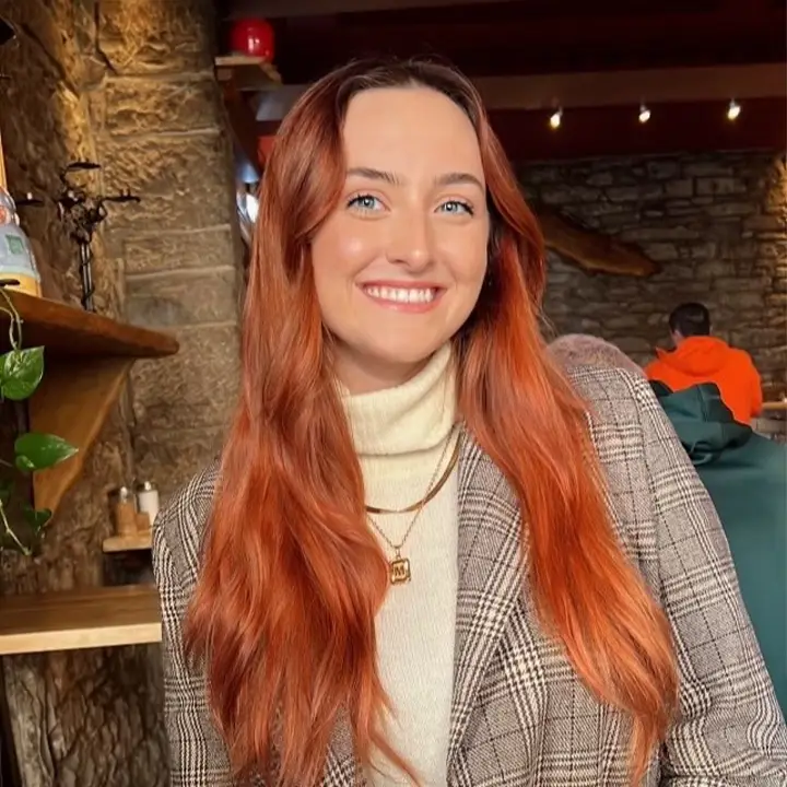 Headshot of a smiling young woman with long red hair and a chequered jacket