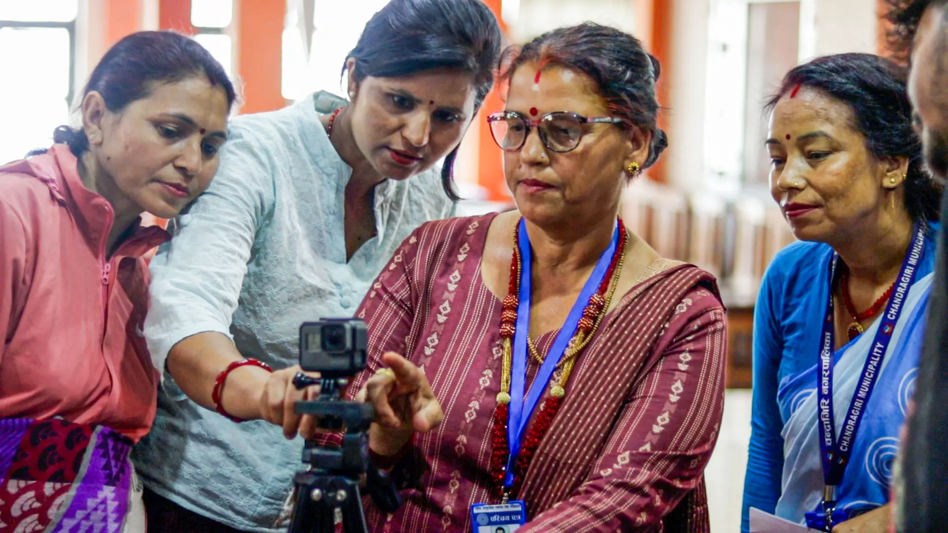 Five Nepali women stand examining a camera on a tripod