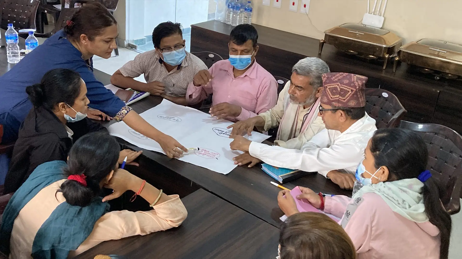 Workshop in Lumbini Province, Nepal showing a standing woman in a blue dress pointing to a diagram on a large sheet of paper on a table while men and women sit around