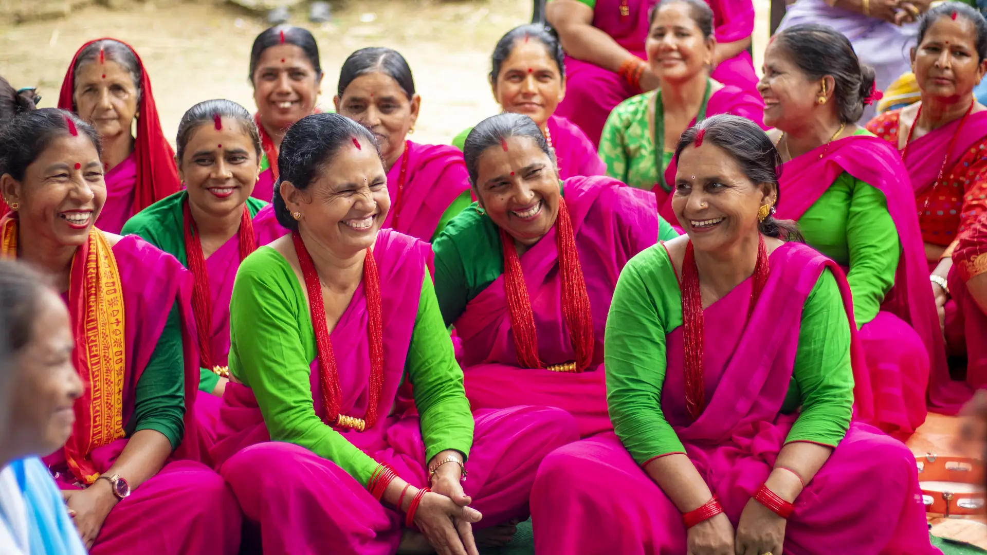 Lots of smiling Nepali women in pink and green saris sit smiling and laughing