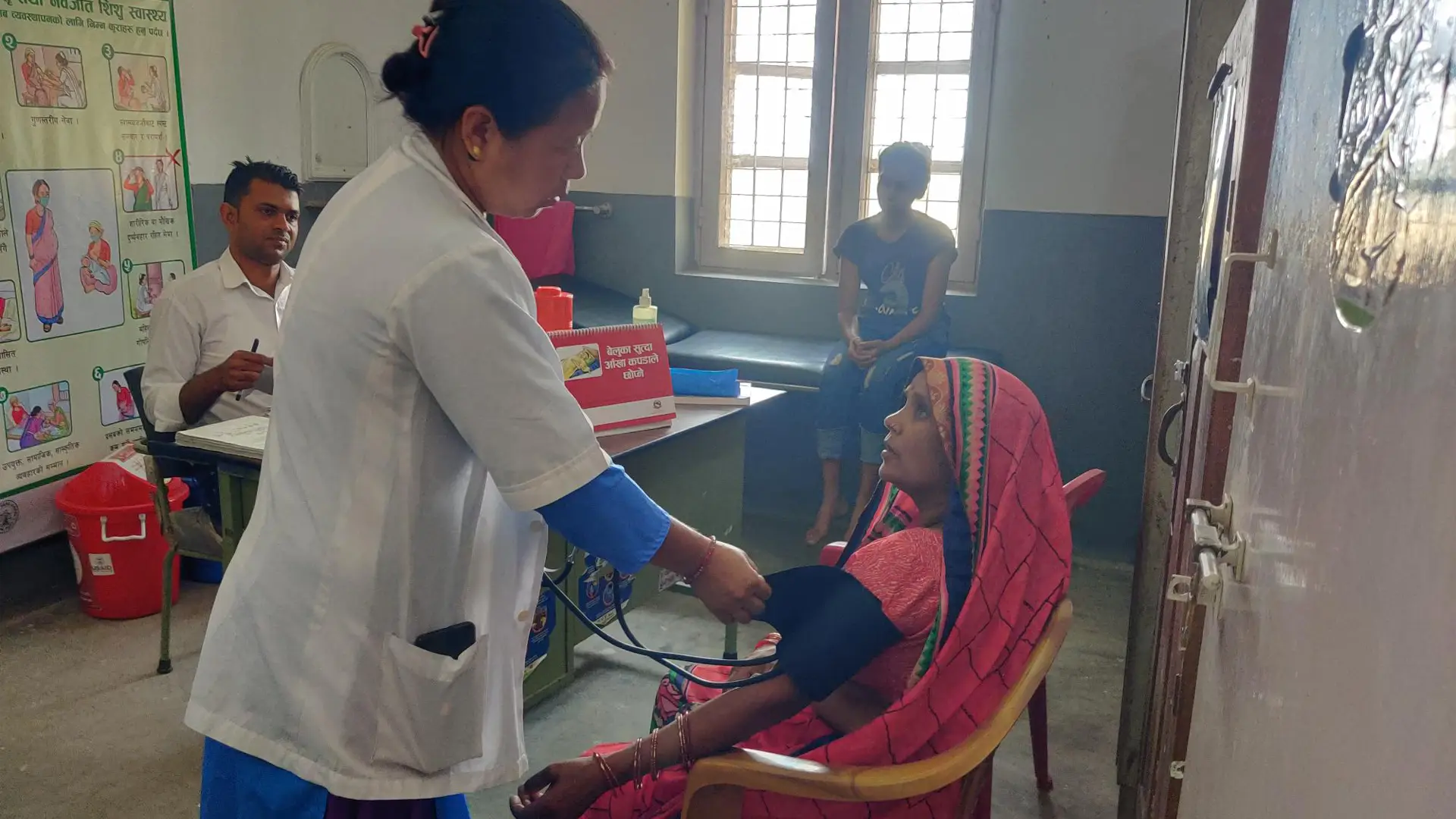 Female doctor in white coat stands taking the blood pressure of a seated Nepali woman in a pink sari