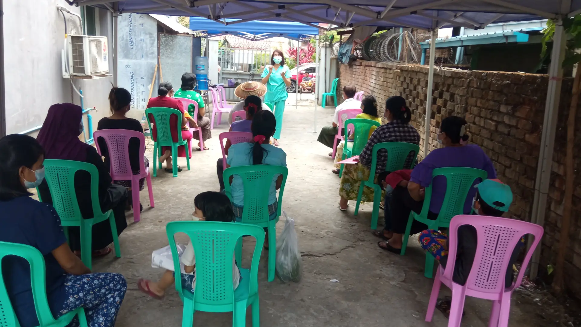 Rows of Burmese people sitting in green and pink chairs while a standing woman wearing a mask speaks to them