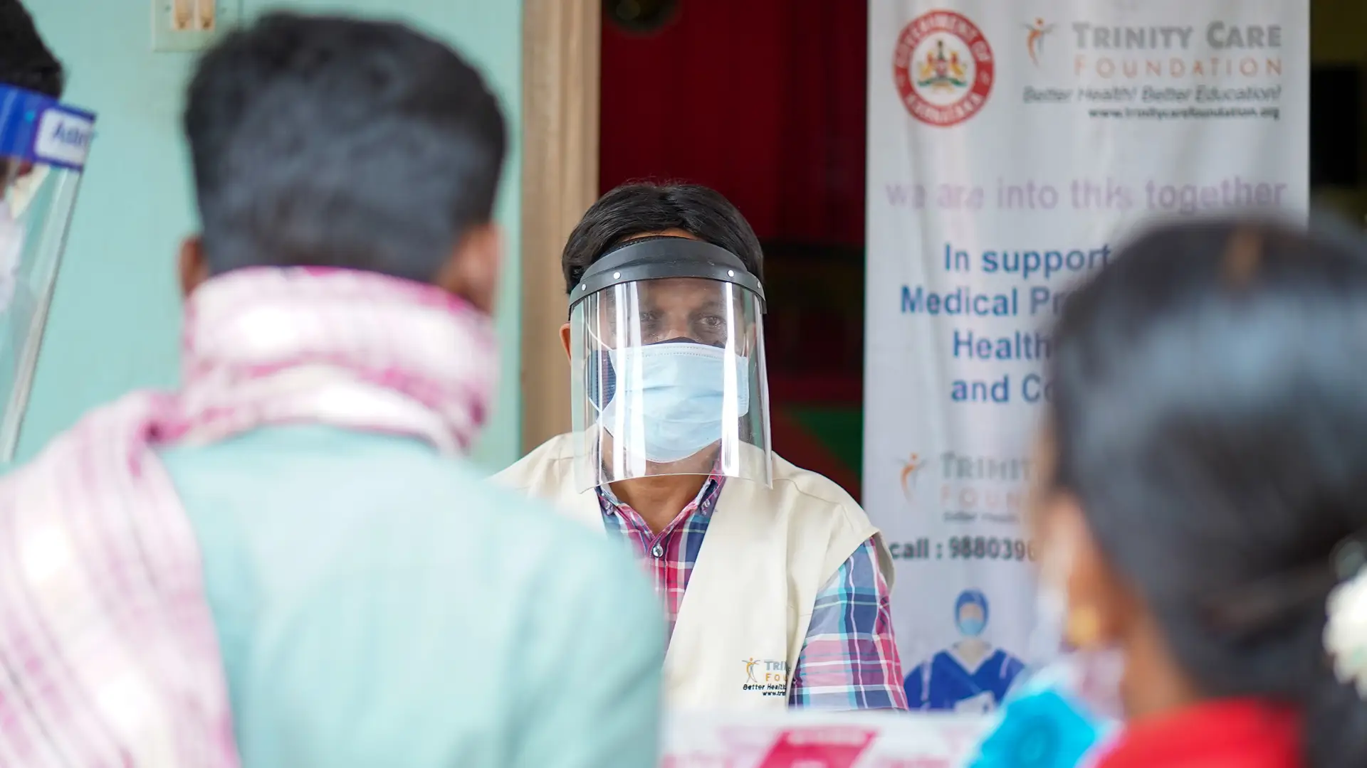 An Indian man in face mask and face guard speaks to a couple opposite him