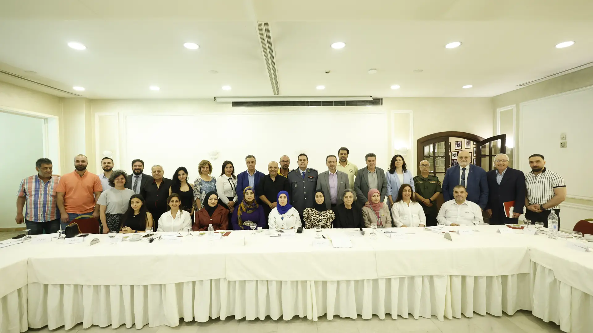 A group of about 30 Syrian and Lebanese men and women sit and stand at table, looking at the camera. The walls and table are white.