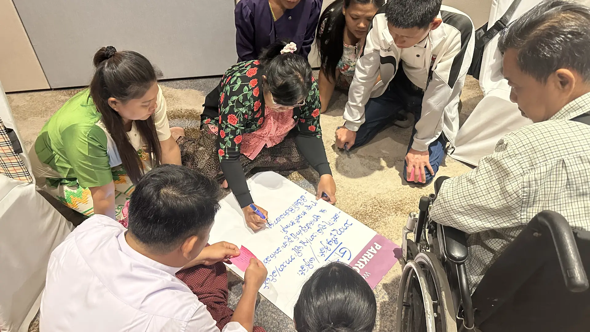 A group of Burmese people sit on the floor around a sheet of flip chart paper. A woman writing on the paper. One of the men is sitting in a wheelchair.