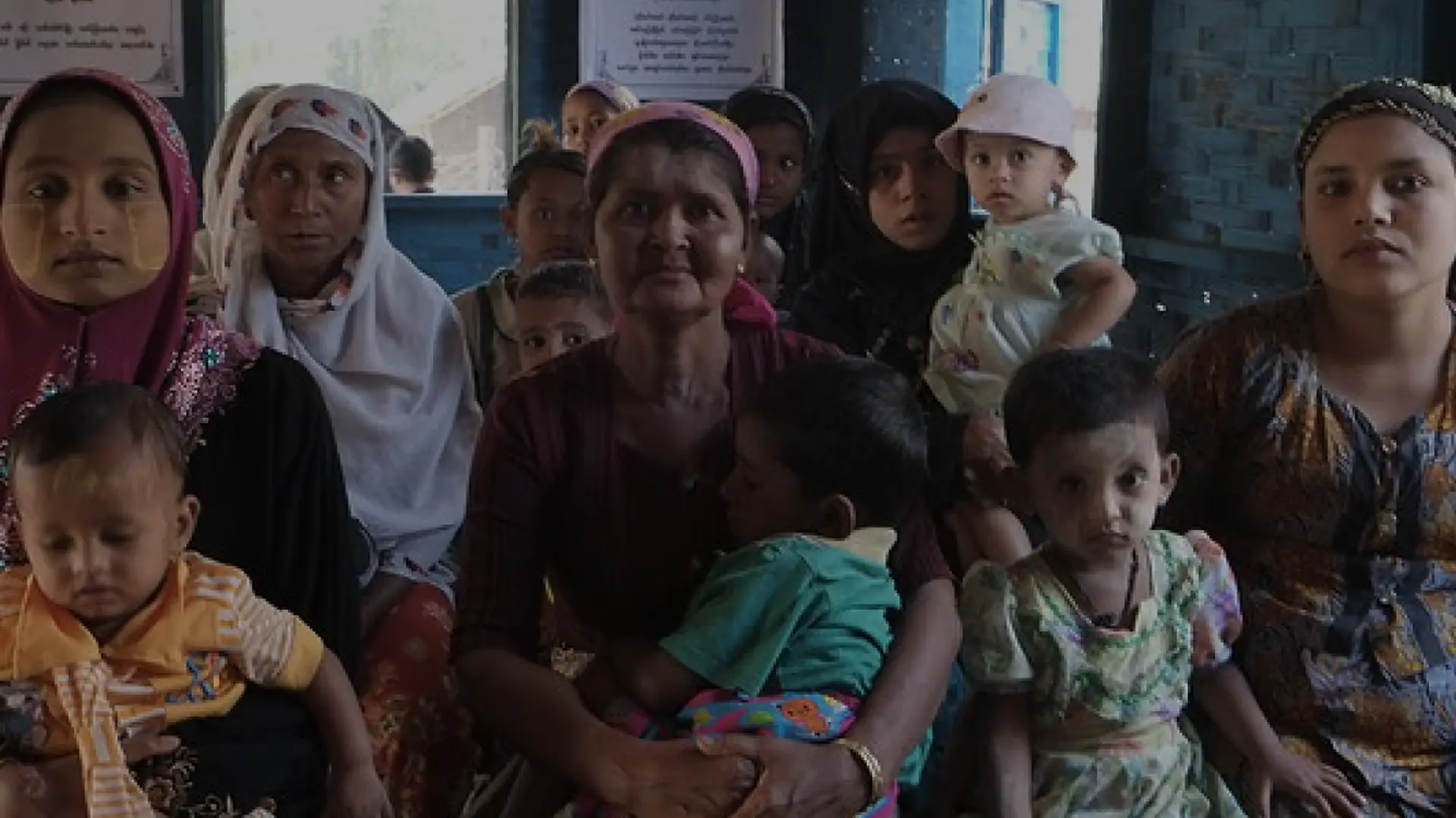 Photograph taken in a health clinic waiting room in Bar Sar Yar camp for displaced people in Rathedaung Township, Myanmar. Rohingya women and children sit looking at the viewer