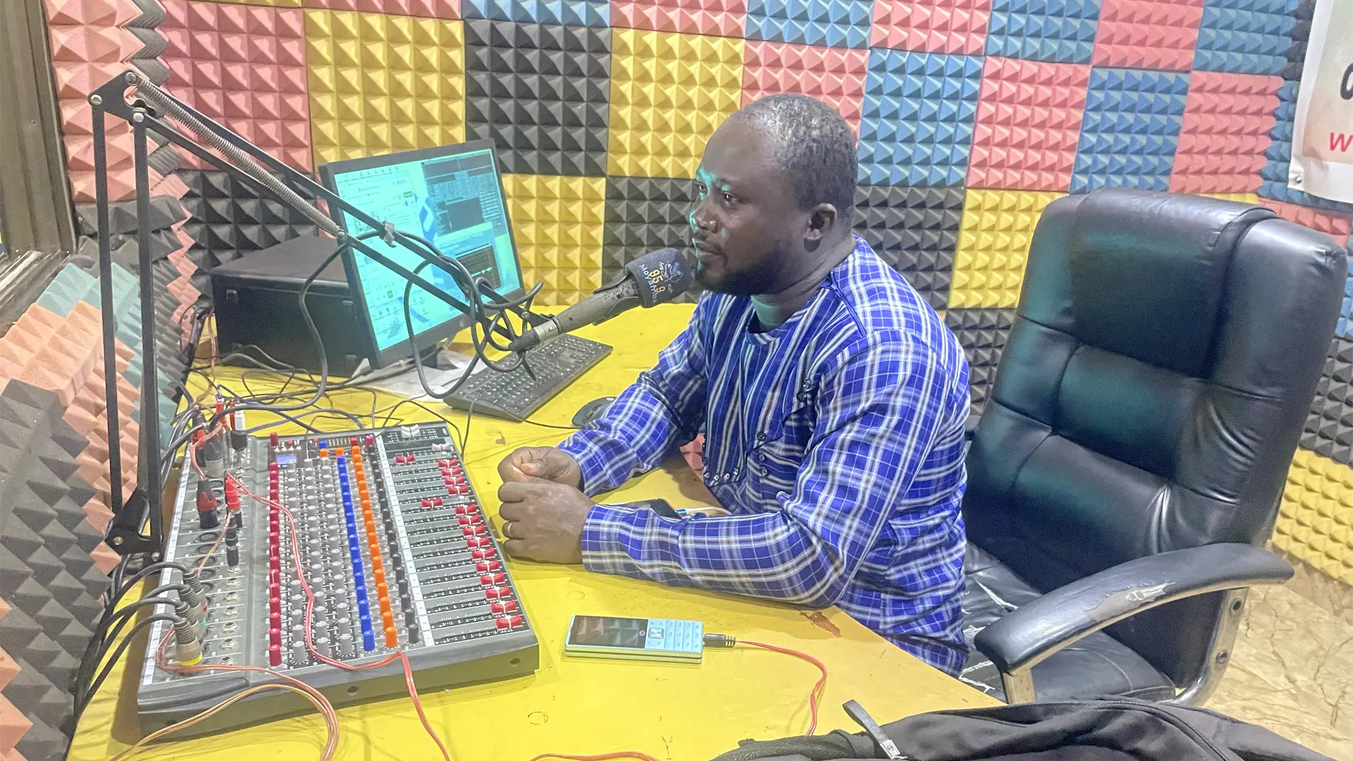 It's a radio studio with brightly coloured walls and a Sierra Leonian man speaking into a microphone in front of a mixing desk
