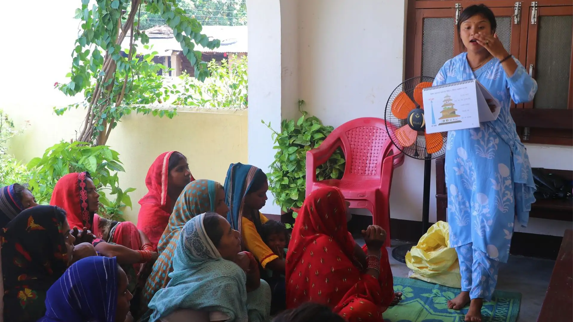 A female community health volunteers stands with a diagram, speaking to seated women and children