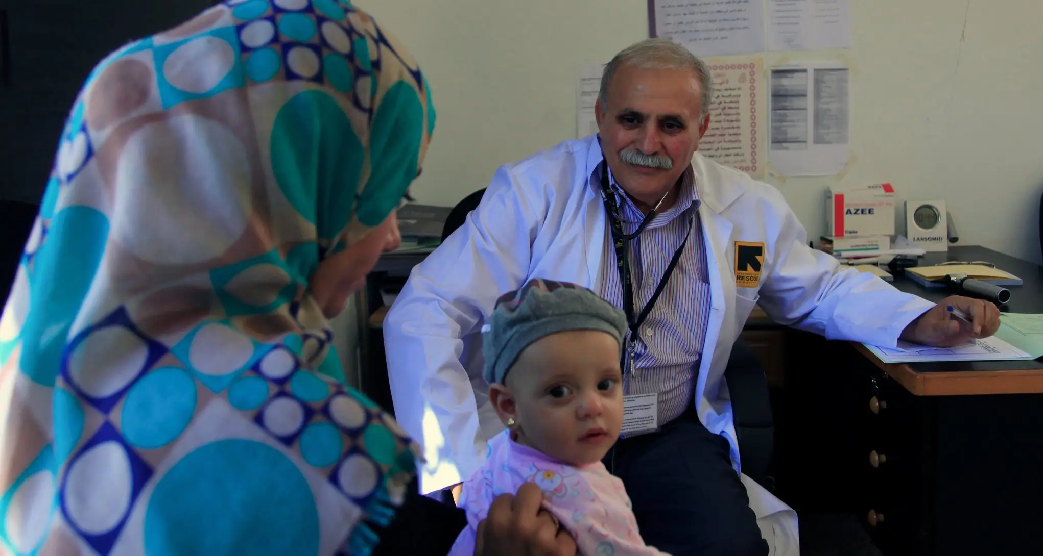 A male IRC doctor in a white coat smiles at a child on its mother's knee - the child is turning to look at the viewer