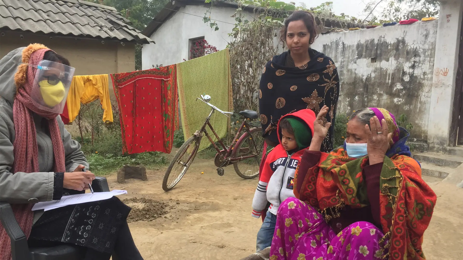A Nepali woman in a plastic mask interviews an older woman while watched by a younger woman and child