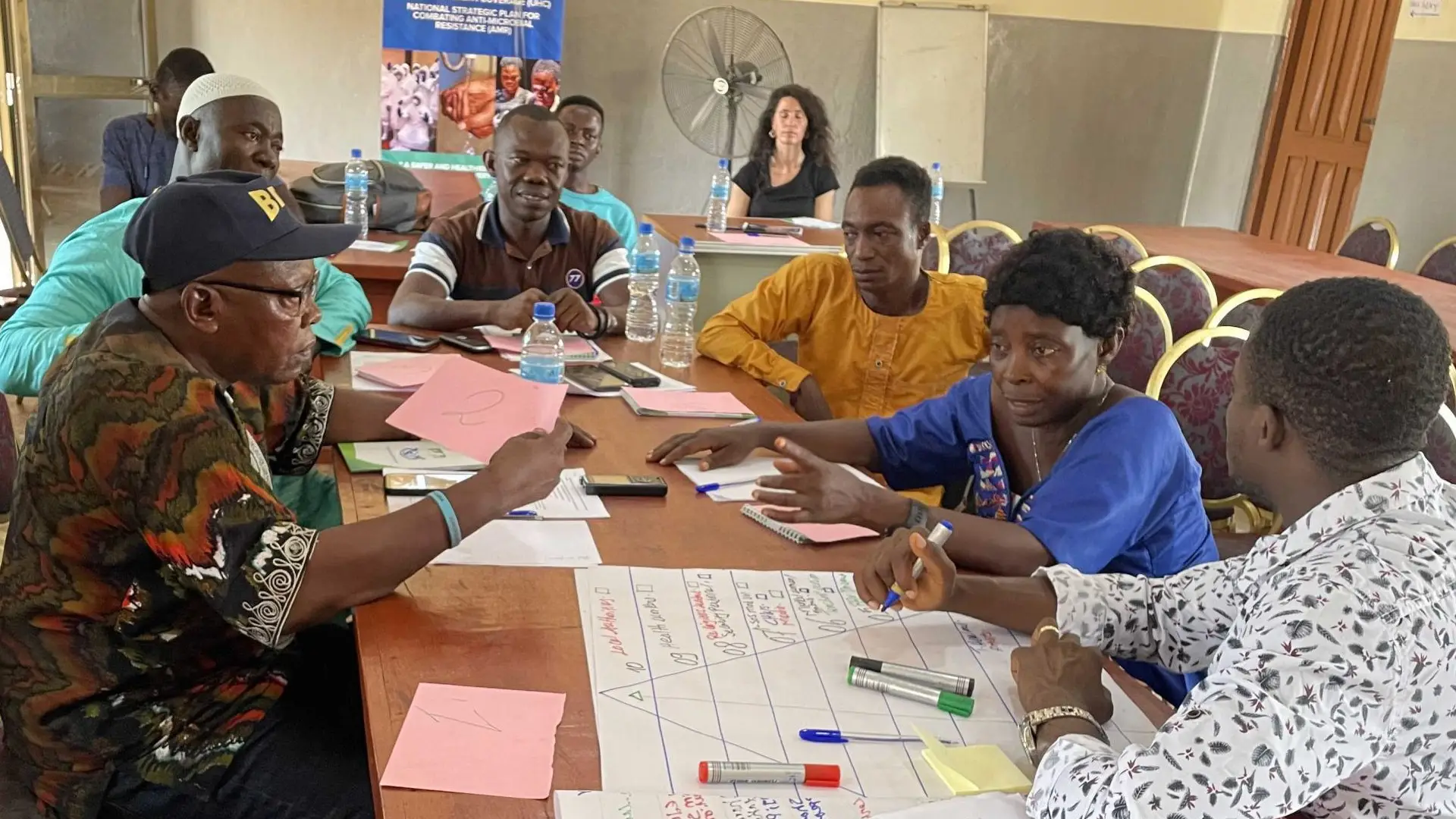 A group of Sierra Leonian men and women sit talking around a table - they are drawing a diagram on flipchart paper