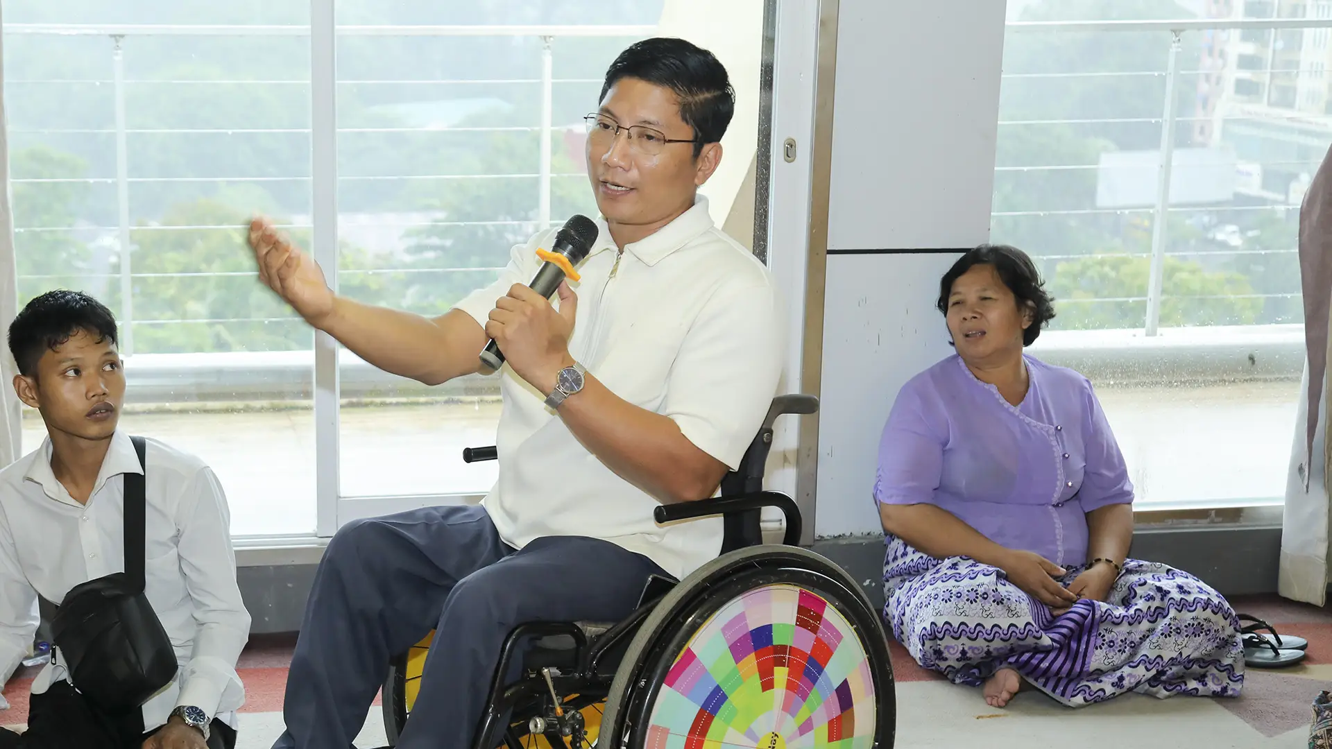 A Burmese man in a wheelchair speaks into a microphone while gesticulating and being watched by people sitting around him