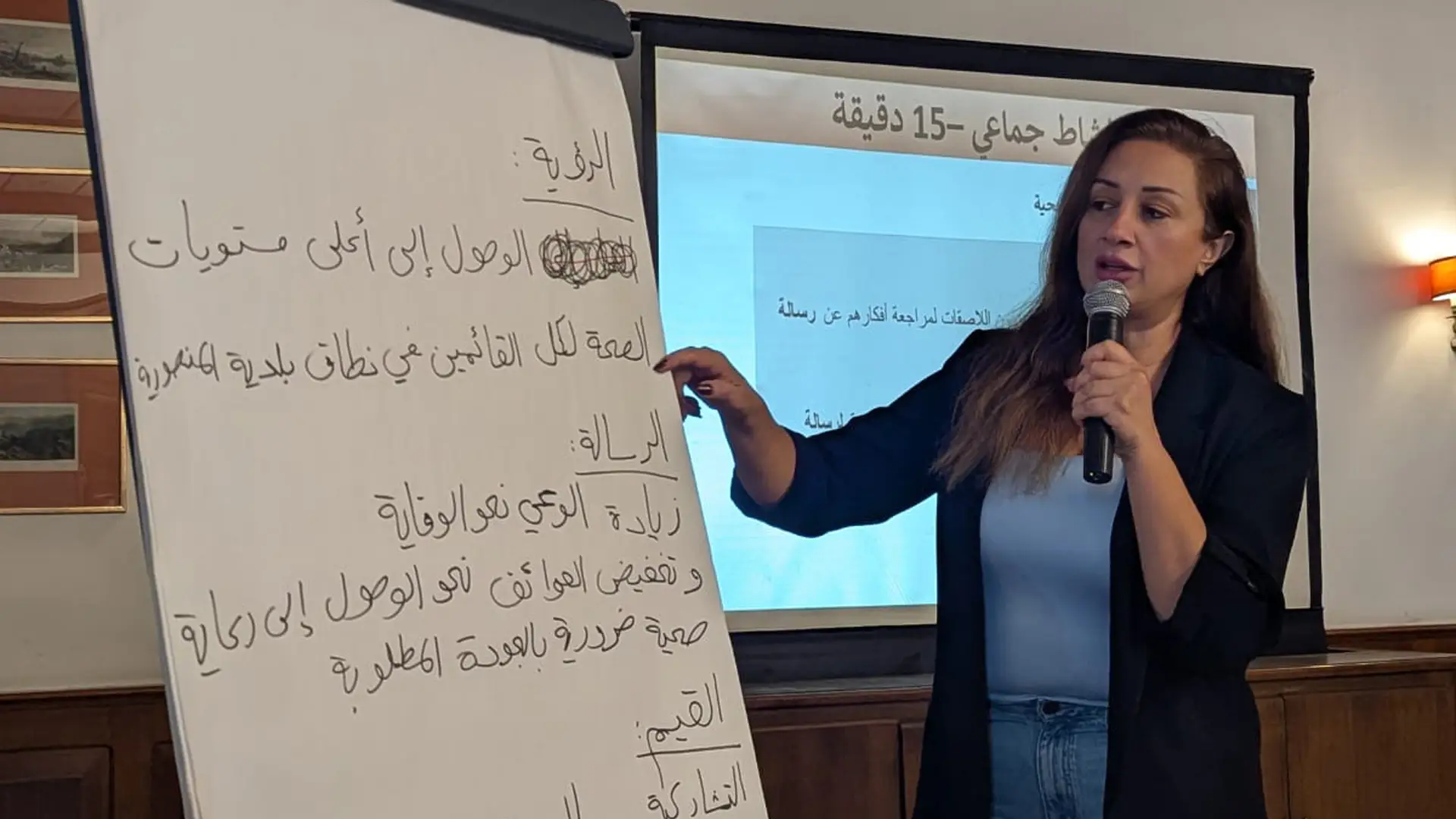Lebanese woman points with a sheet of flip chart paper with Arabic writing. She's speaking into a microphone
