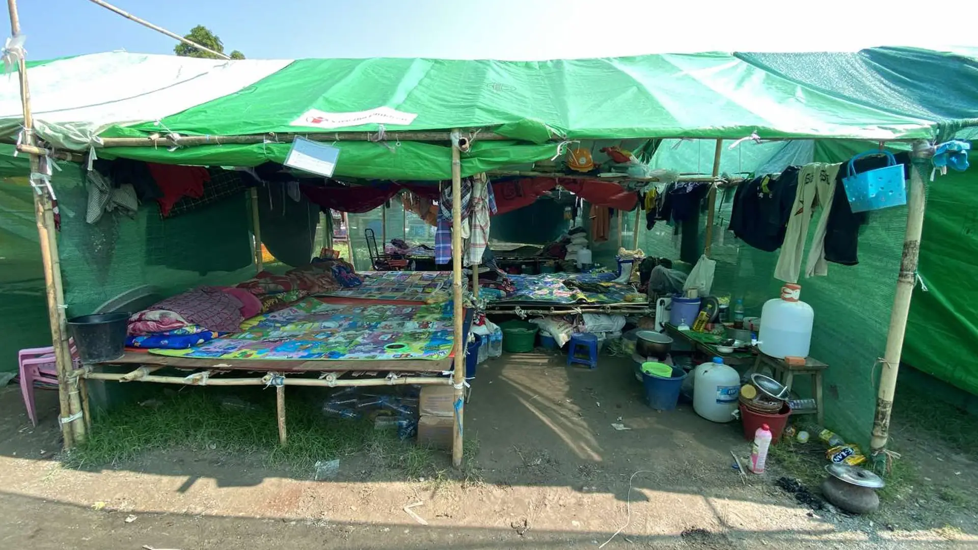 A bamboo shelter with green tarpaulin. Inside are three beds, hanging clothes and buckets and bottles of water